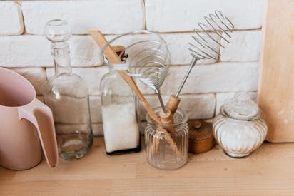 A collection of glass jars and kitchen utensils on a rustic wooden counter with a white brick background.