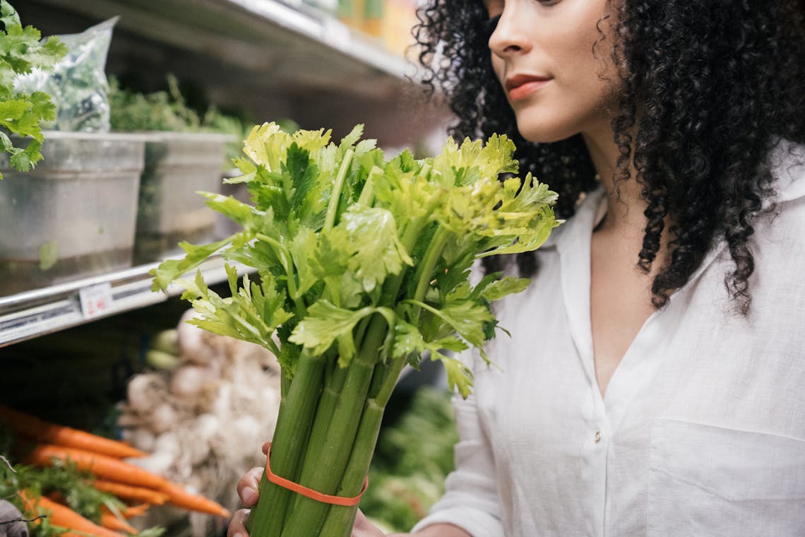 A woman examines a fresh bunch of celery in a produce aisle at the grocery store.